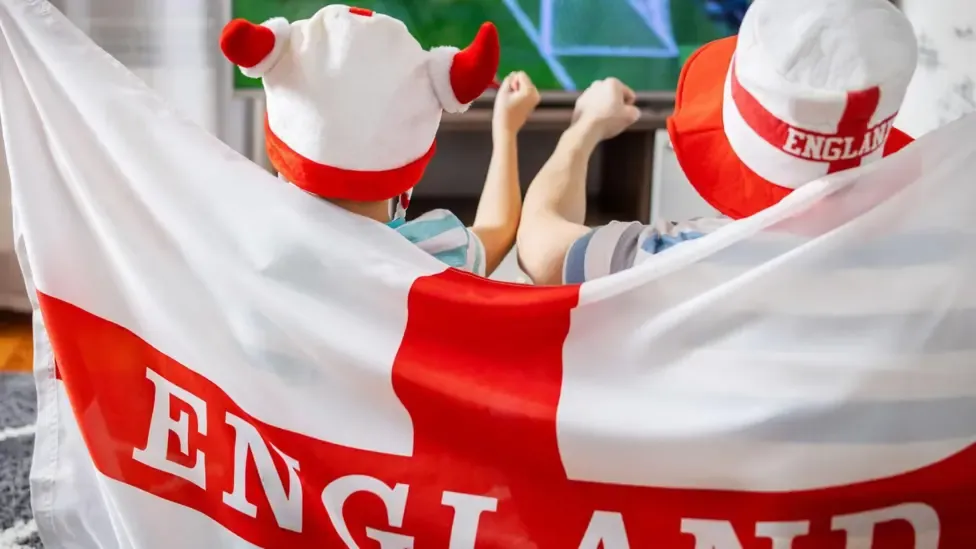 Two young England fans drapped in a ST George's cross cheer as they watch the Three Lions play on tv.